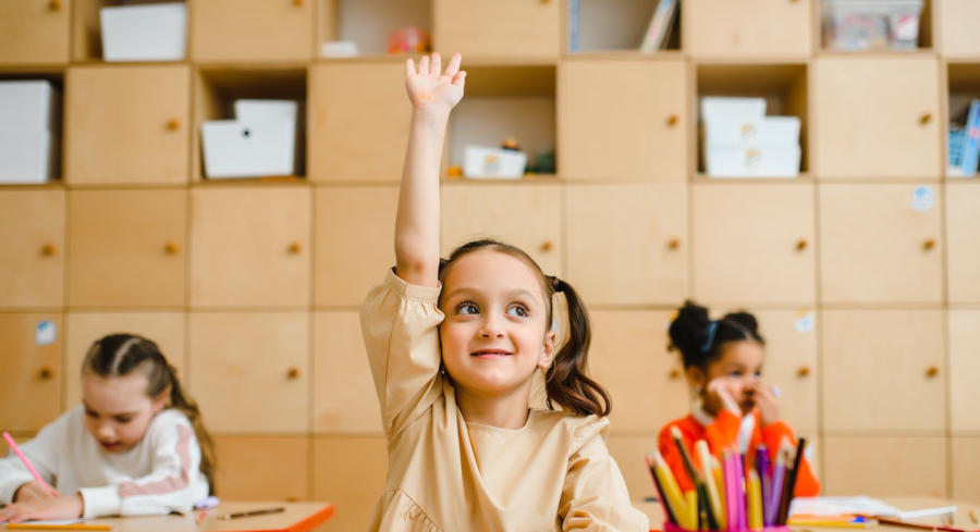A girl in the center of two girls raising her hand in a classroom