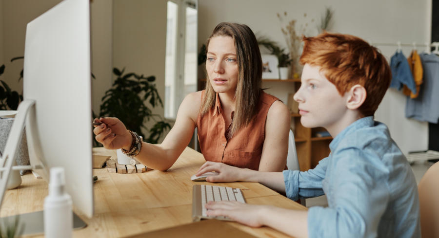 A mother pointing at a computer monitor screen, reviewing information with a child sitting next to her